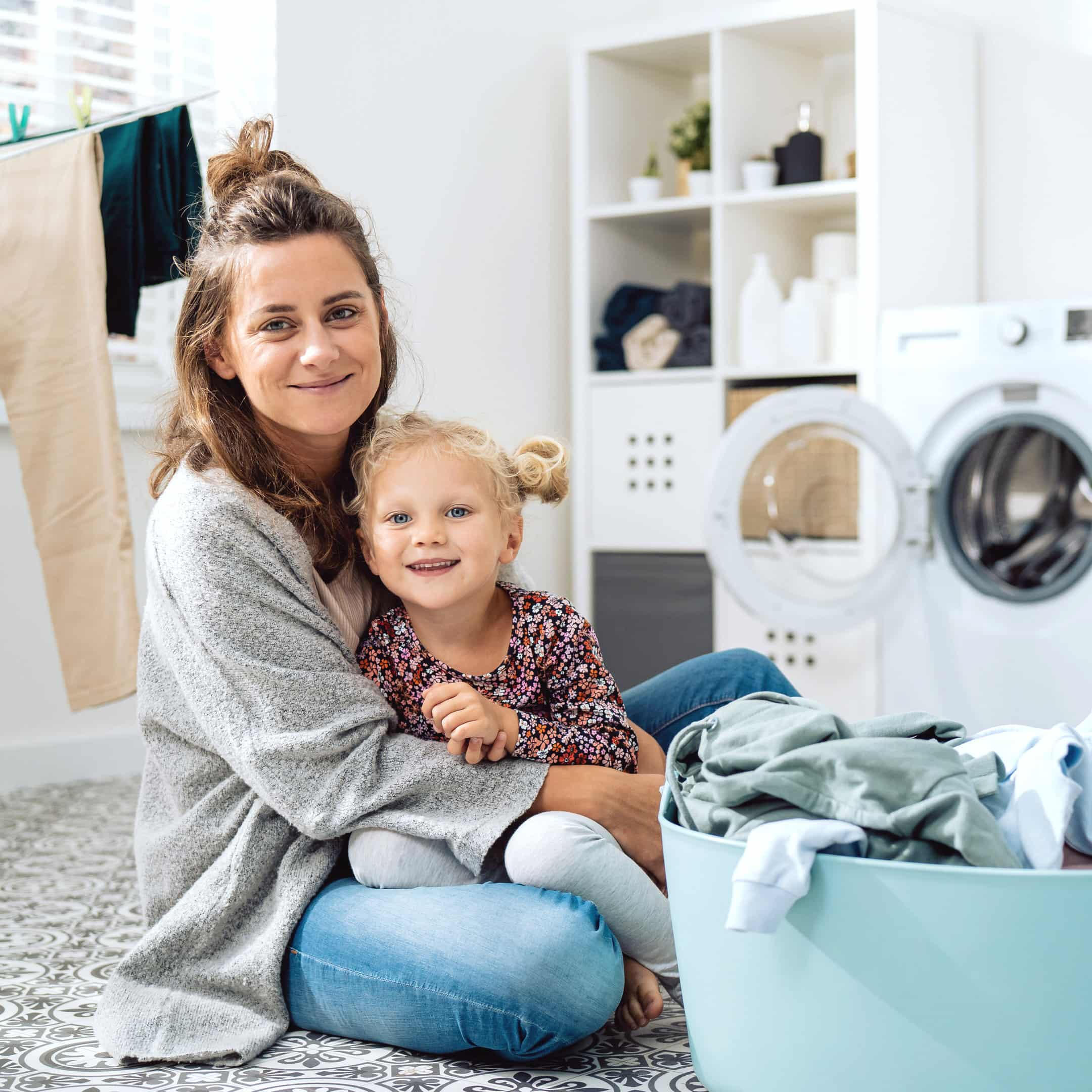 Mother and child sitting in a laundry room with a basket of clothes, representing Viola Cleaning Journal’s practical home cleaning guidance